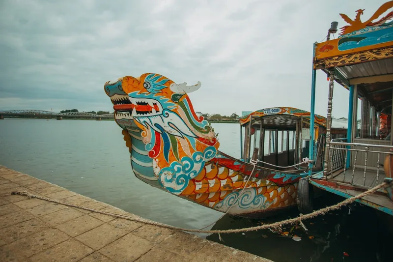 A colorful carved dragon boat is docked along a riverfront under a cloudy sky in Vietnam.