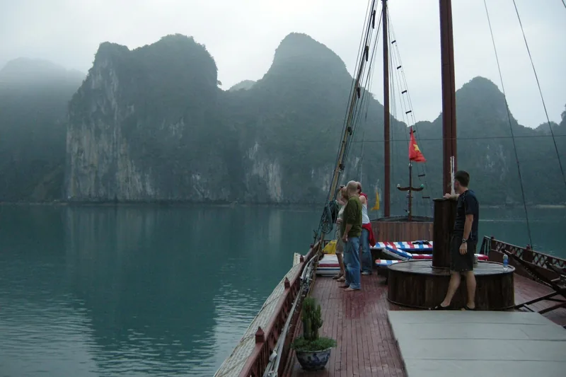 Passengers on a wooden junk boat deck admire misty limestone karsts in Ha Long Bay.