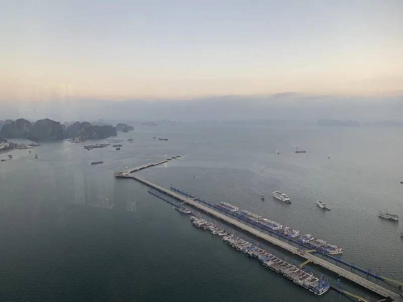 Aerial view of a long pier with moored tour boats and misty limestone islands in Ha Long Bay at dusk.