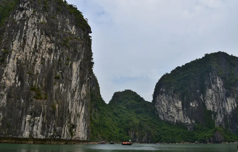 Towering limestone karsts rise above the calm water of Ha Long Bay with small boats passing between them.