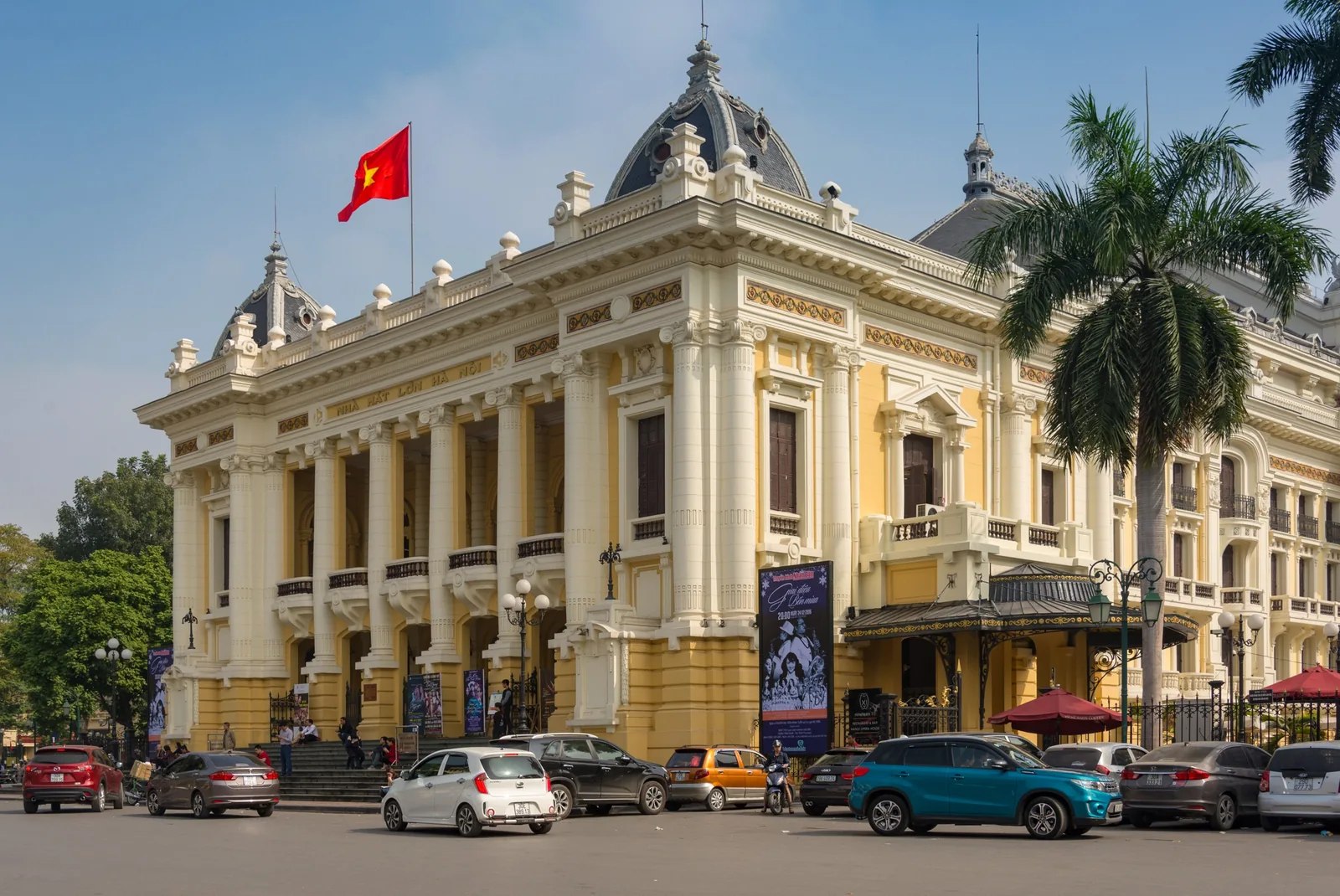 The French colonial facade of the Hanoi Opera House rises above parked cars under a clear blue sky.