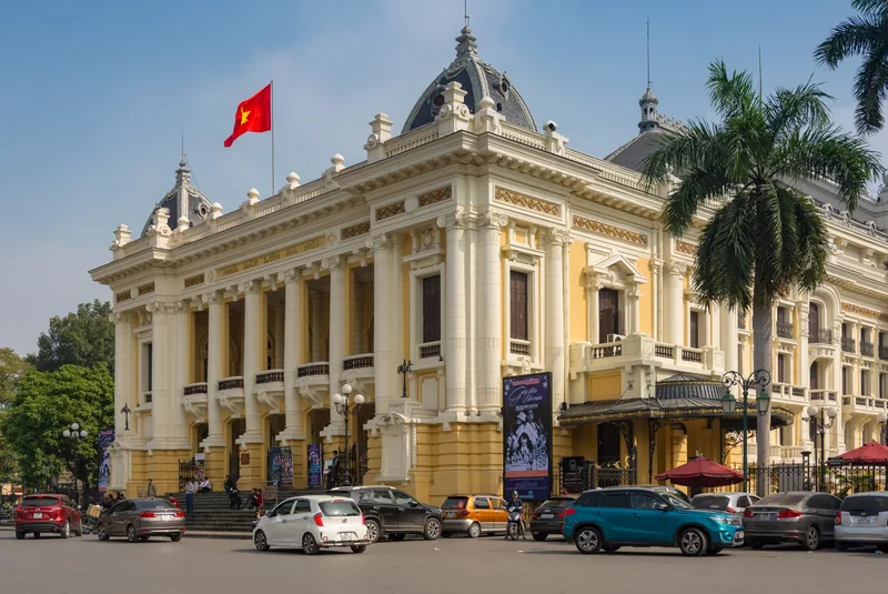 The French colonial facade of the Hanoi Opera House rises above parked cars under a clear blue sky.