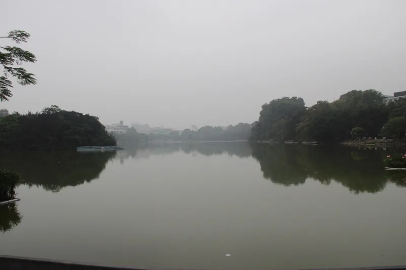 Misty morning view across Hoan Kiem Lake in Hanoi, with tree-lined shores and calm reflective water.