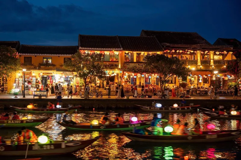 Lantern-lit boats drift along the Thu Bon River beside glowing riverside houses in Hoi An at night.