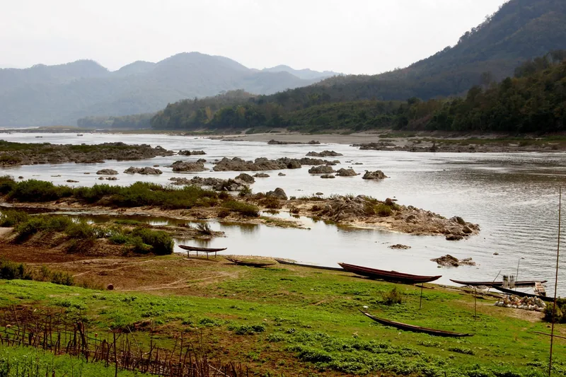 Wooden long boats rest on the banks of the Mekong River near Luang Prabang, framed by misty mountains.