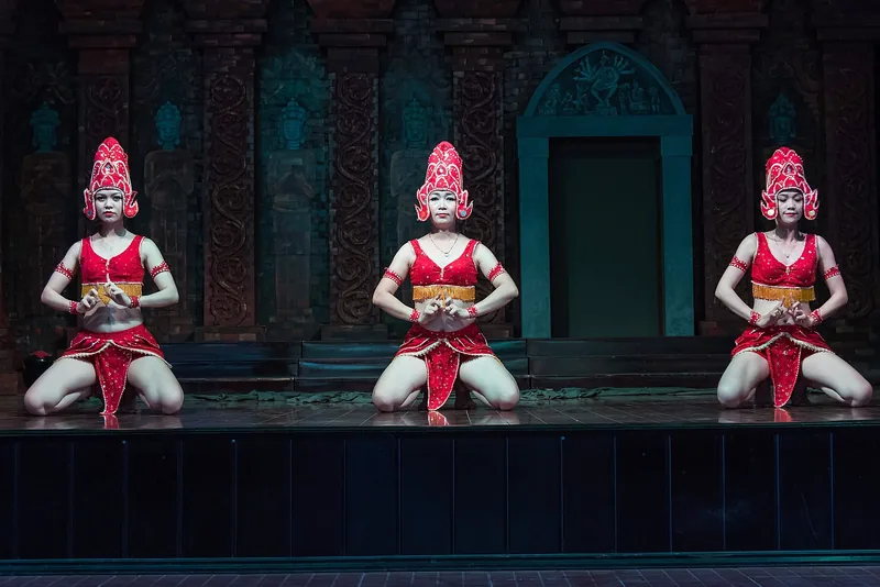 Three dancers in red Cham costumes and headdresses perform kneeling on stage against a carved temple backdrop at My Son.
