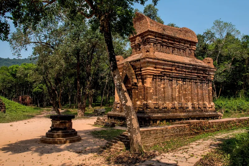 A weathered brick tower of My Son sanctuary stands amid dense forest under a clear blue sky.