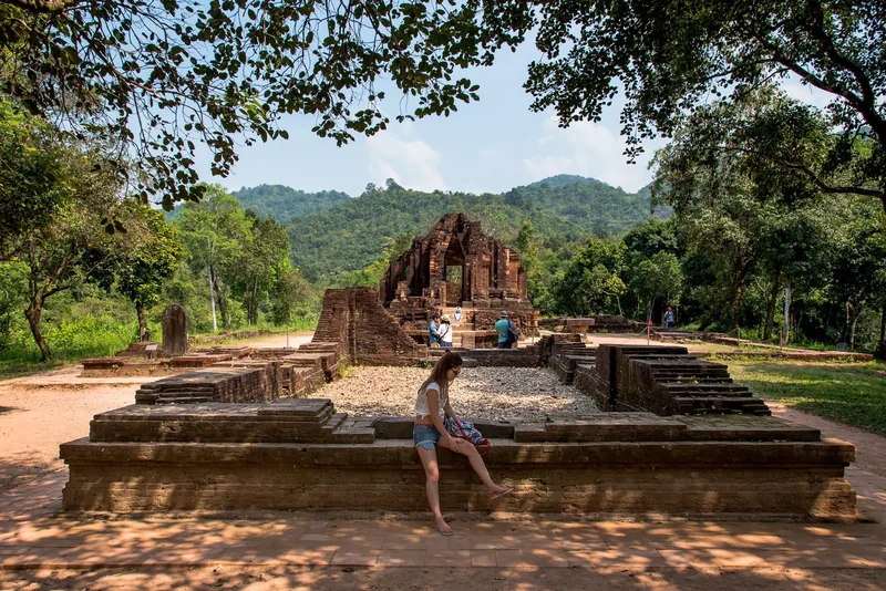 A visitor sits on ancient brick ruins at My Son Sanctuary, with forested hills rising behind the temple towers.