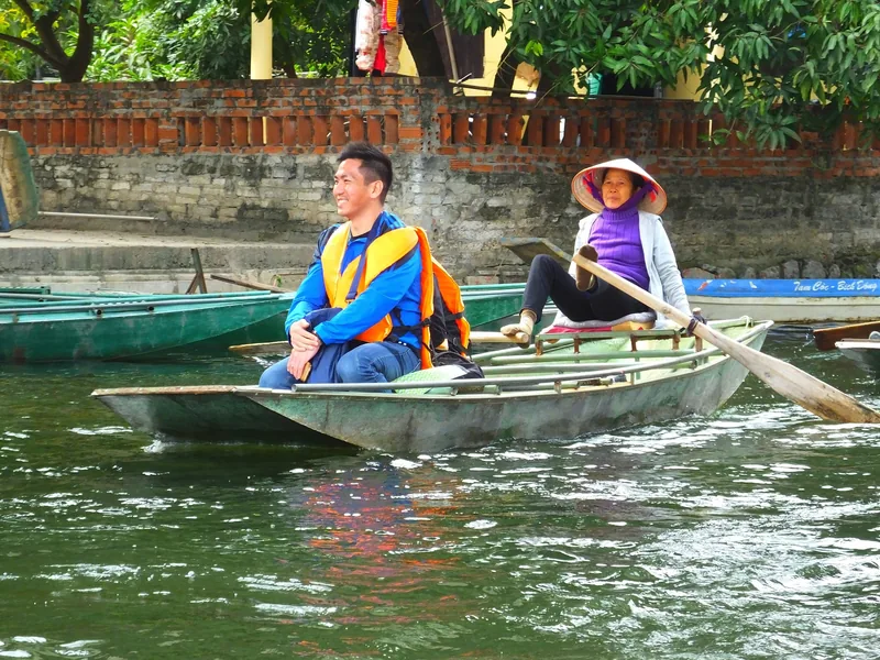A woman in a conical hat rows a sampan with her feet while a tourist rides along a river in Ninh Binh