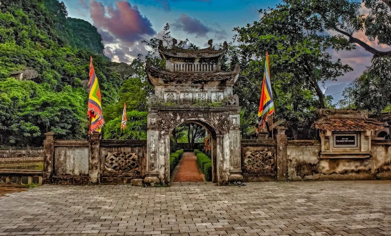 Ancient stone gate with colorful flags at Hoa Lu ancient capital in Ninh Binh, surrounded by limestone hills.