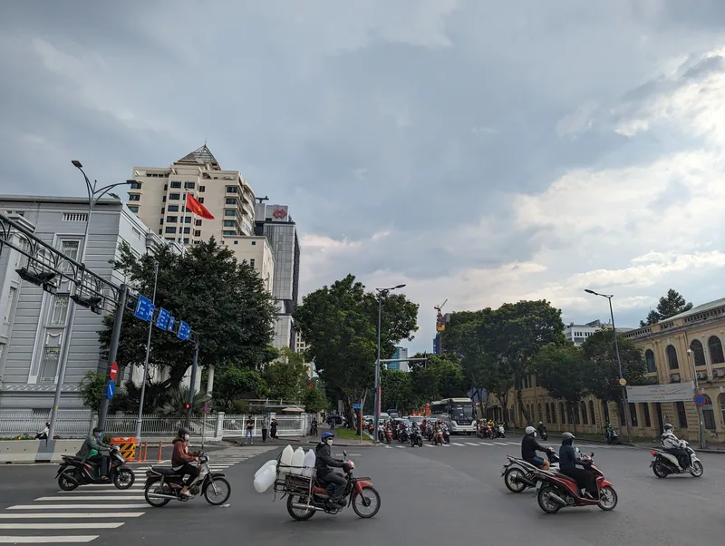 Motorbikes cross a wide intersection lined with trees and colonial buildings in Ho Chi Minh City.