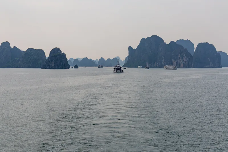 Cruise boats sail among limestone karst islands on Ha Long Bay under a hazy sky.